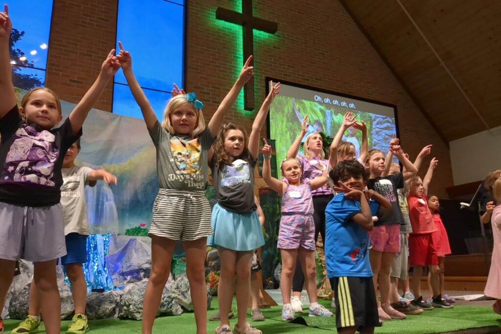 Kids on stage with their arms raised, singing at Vacation Bible School